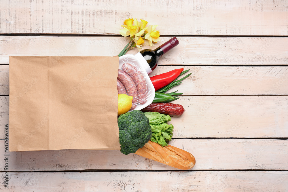 Paper bag with vegetables, sausages and wine bottle on white wooden background