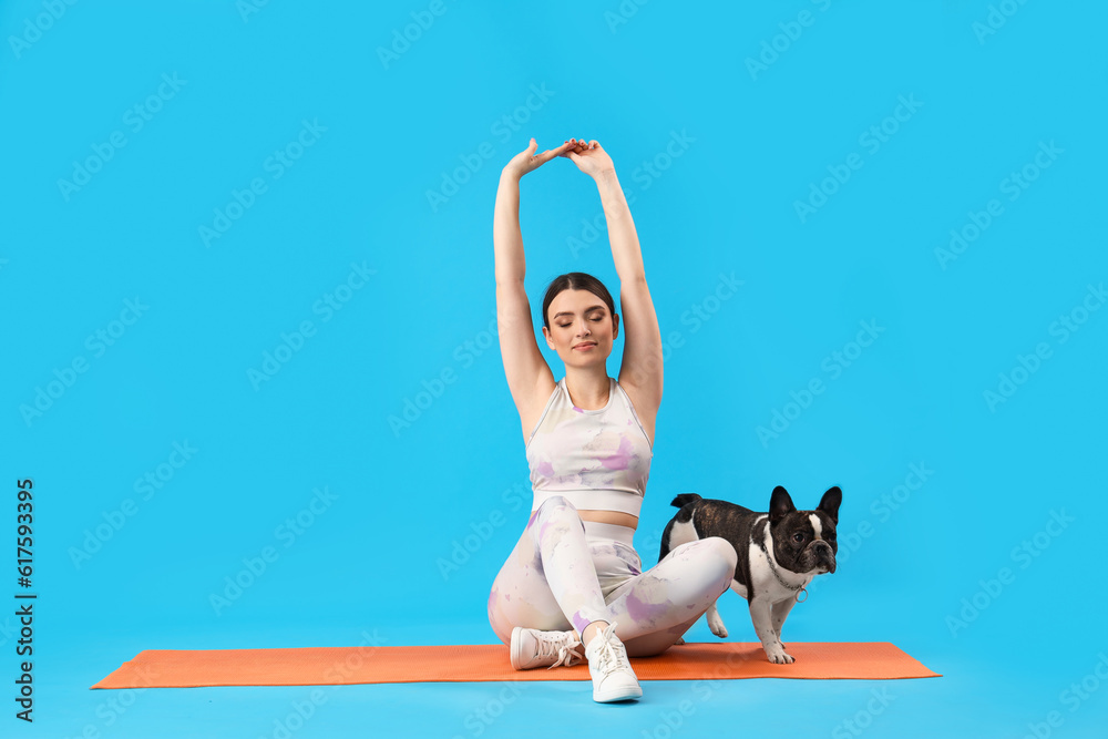 Sporty young woman with her French bulldog stretching on blue background