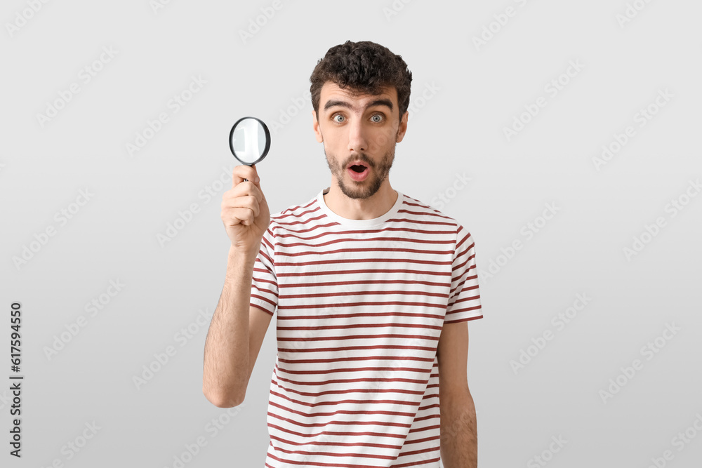 Shocked young man with magnifier on light background