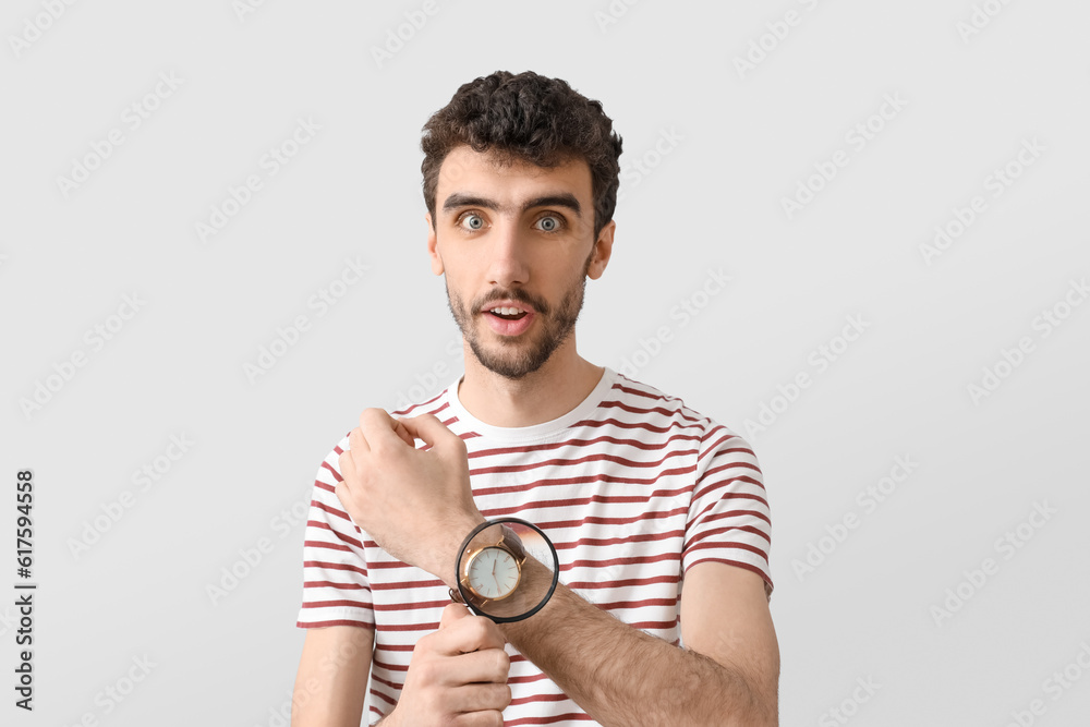 Shocked young man magnifying wristwatch on light background