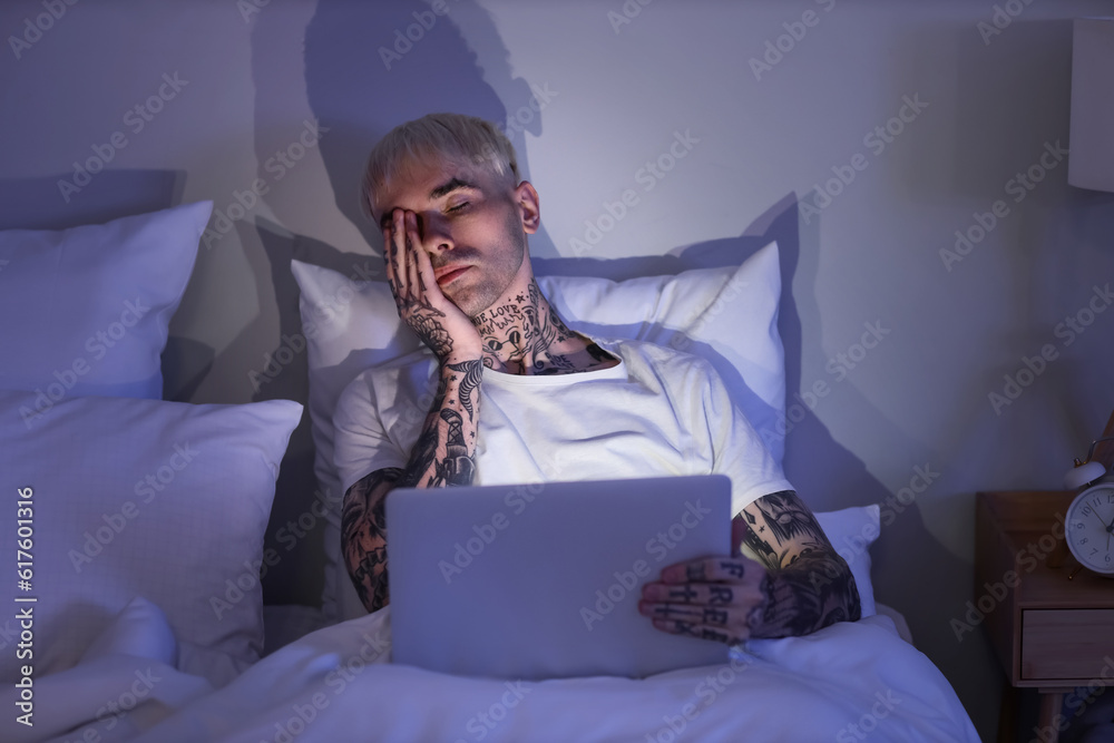 Sleepy young tattooed man using laptop in bed at night