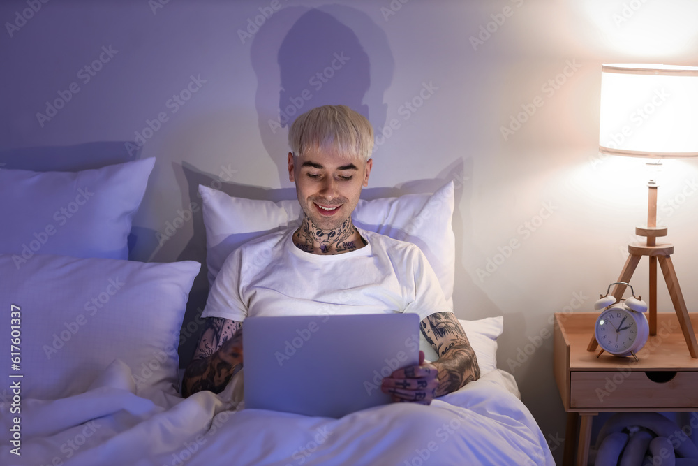 Young tattooed man using laptop in bed at night