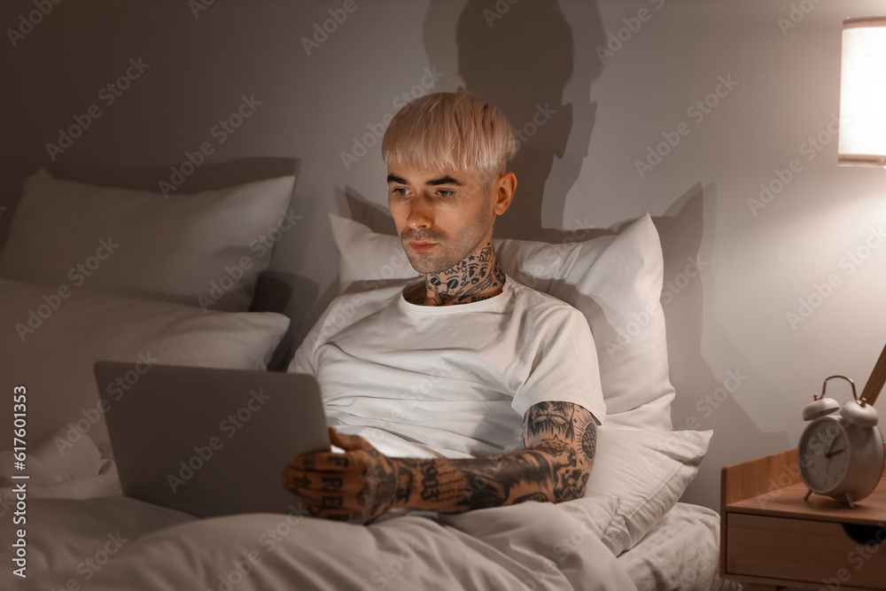 Young tattooed man using laptop in bed at night