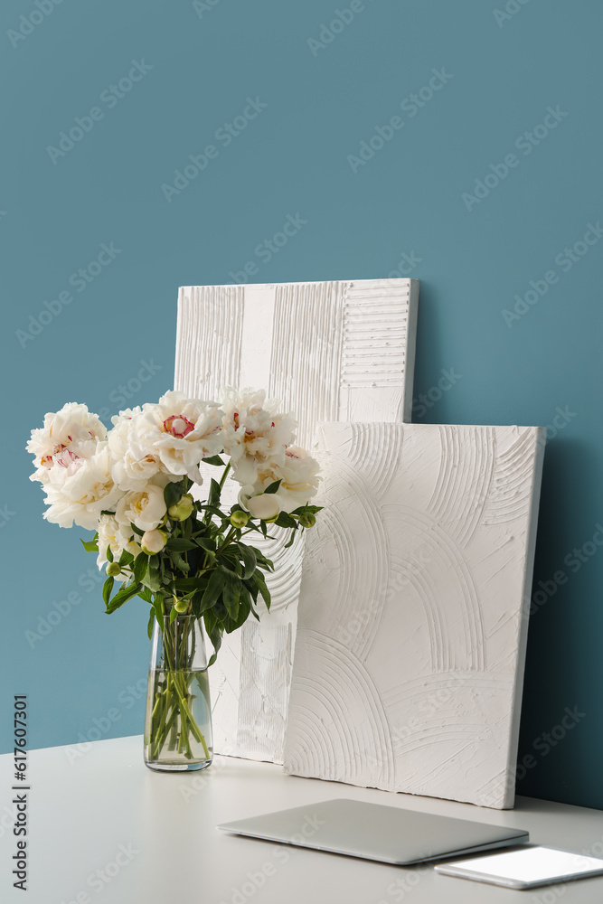 Vase of white peonies and paintings on table near blue wall