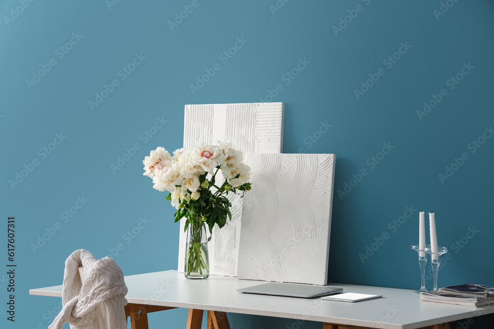 Vase of white peonies and paintings on table near blue wall