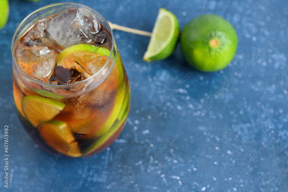 Glass of cold Cuba Libre cocktail and limes on blue table