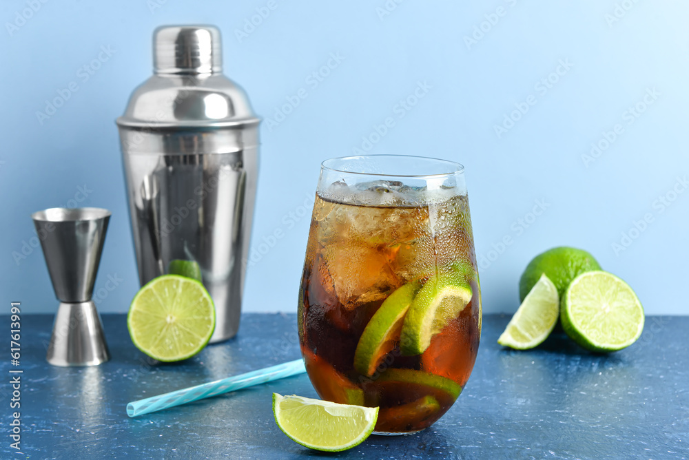 Glass of cold Cuba Libre cocktail and shaker on blue table