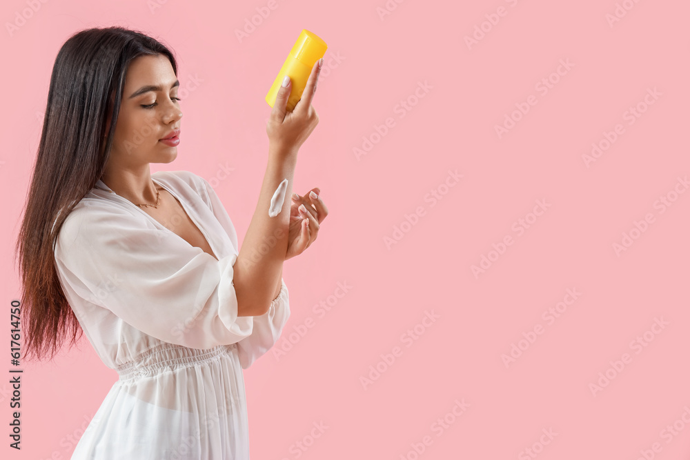 Young woman applying sunscreen cream on pink background