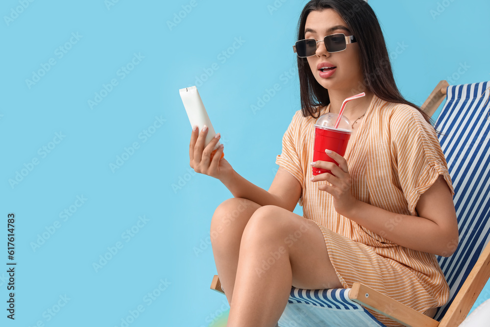 Young woman with sunscreen cream and soda in deck chair on blue background