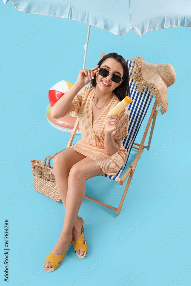 Young woman with sunscreen cream in deck chair on blue background