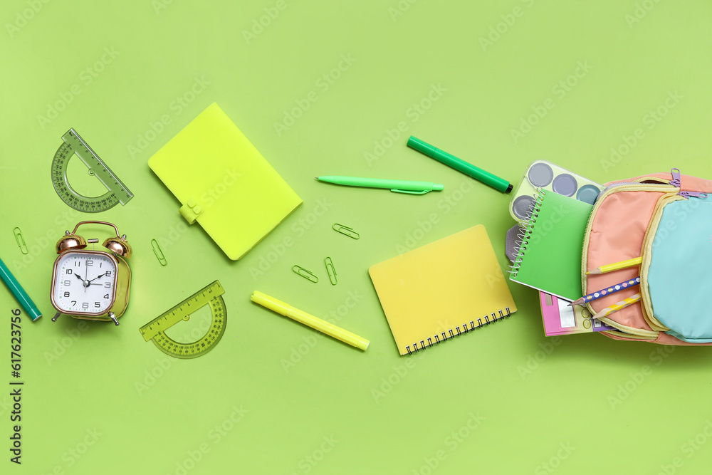 Colorful school backpack with notebooks, alarm clock and pencils on green background