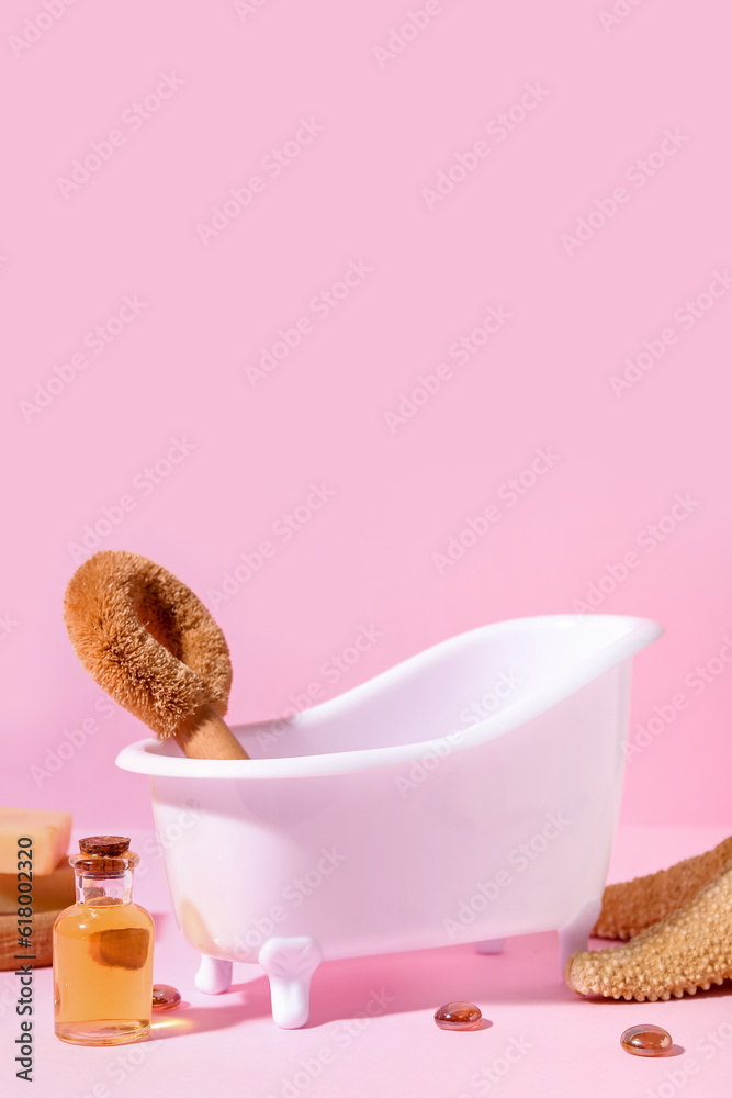 Small bathtub, massage brush and essential oil on pink background