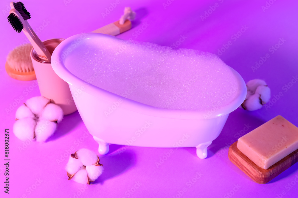 Small bathtub with foam, toothbrushes, soap and cotton flowers on color background