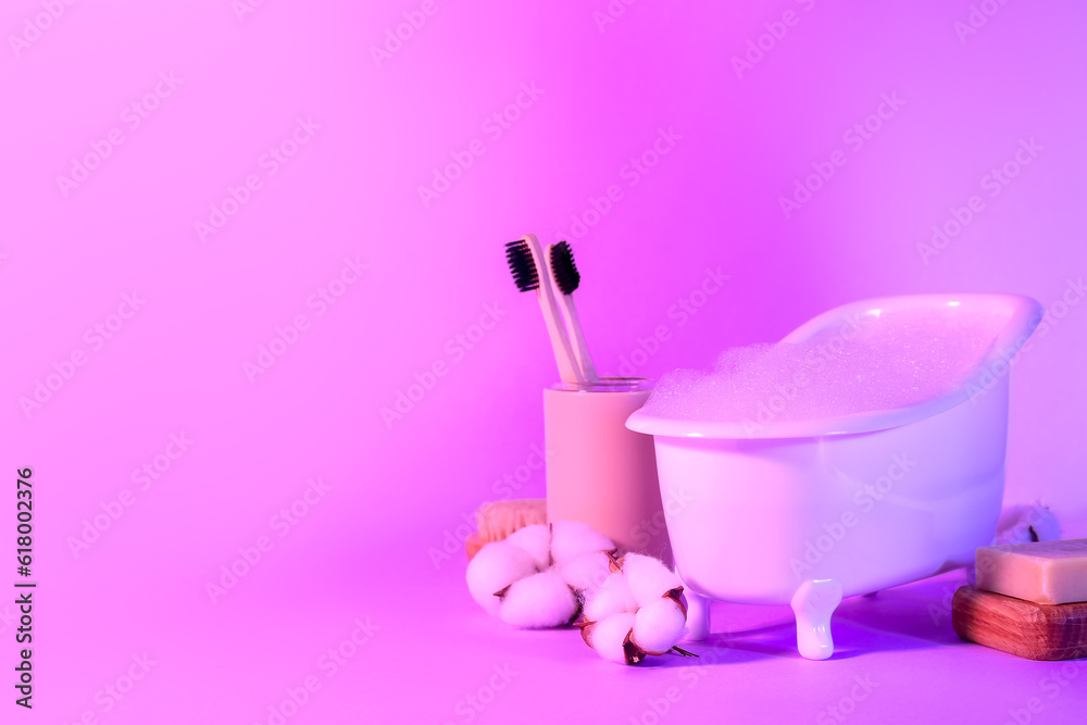 Small bathtub with foam, toothbrushes, soap and cotton flowers on color background