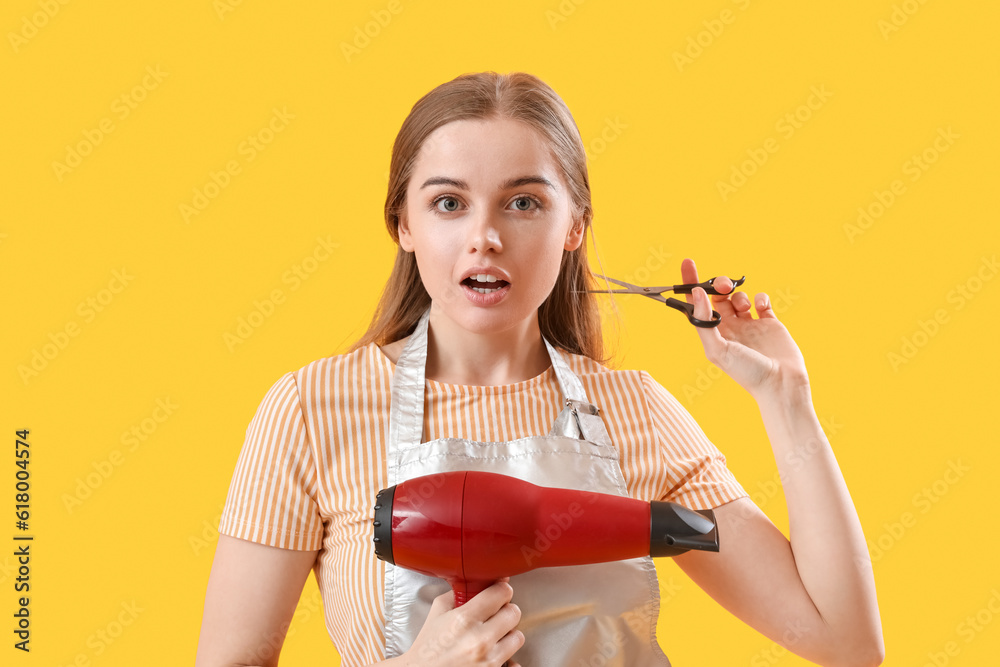 Shocked female hairdresser with dryer and scissors on yellow background, closeup