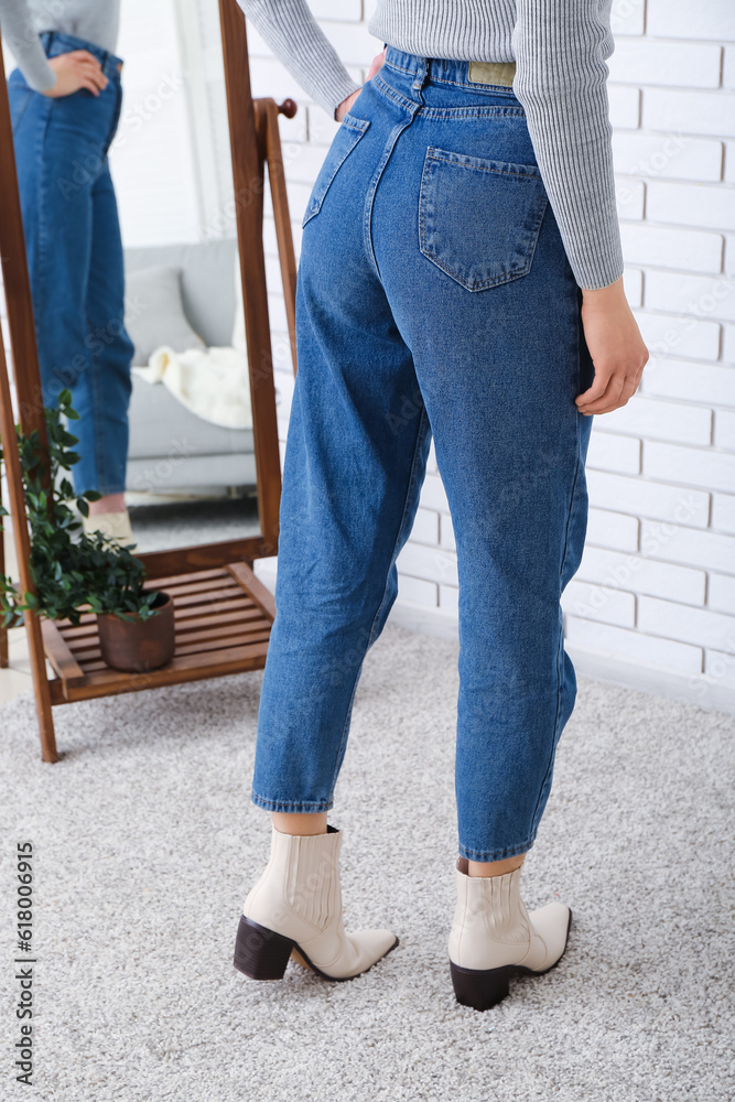 Young woman in stylish jeans and heeled shoes near mirror in room, back view