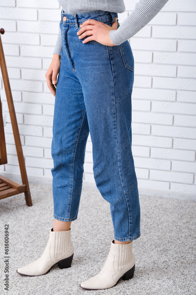 Young woman in stylish jeans and heeled shoes near light brick wall in room