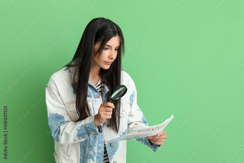 Beautiful young woman with magnifier reading newspaper on green background