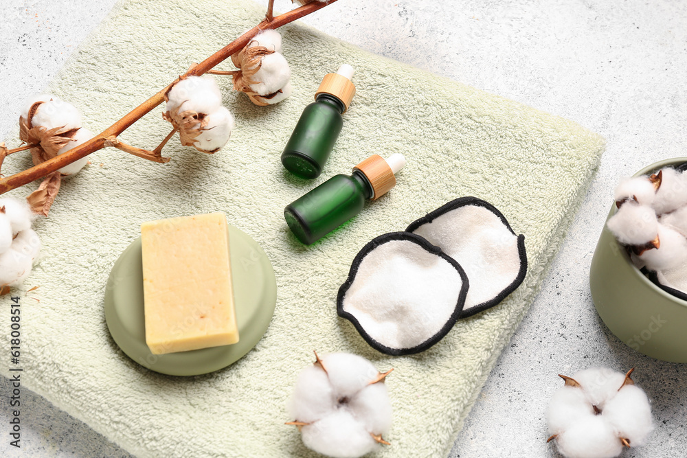 Bottles of essential oil, soap, clean towel, cotton flowers and pads on light background, closeup