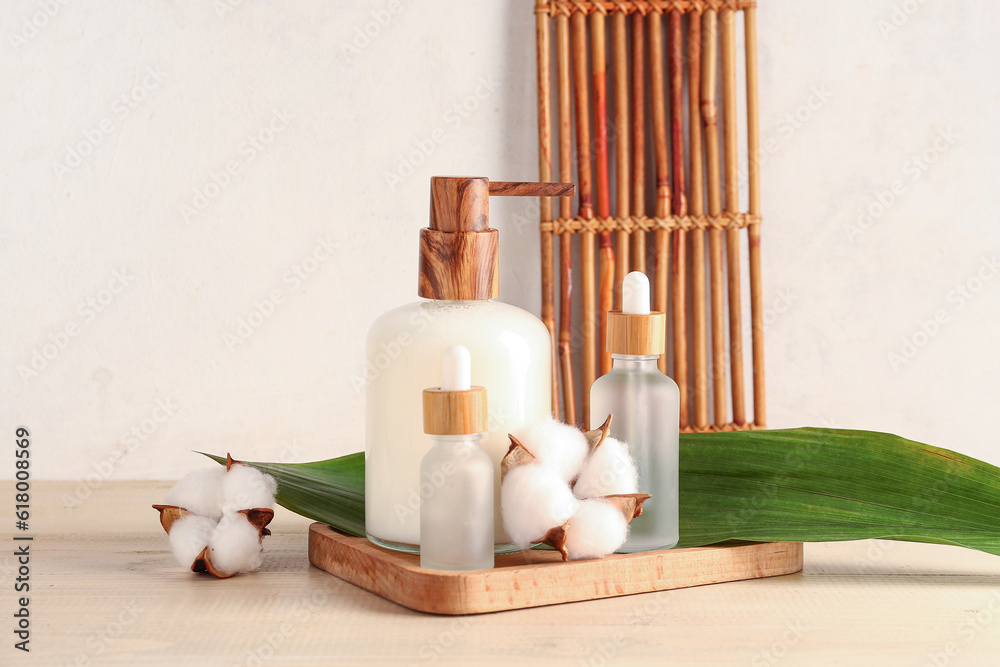 Board with bottles of cosmetic products, cotton flowers and plant leaf on wooden table