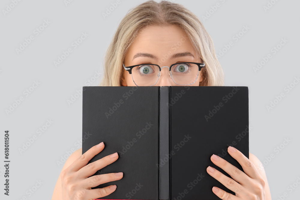 Shocked young woman in stylish eyeglasses reading book on light background, closeup