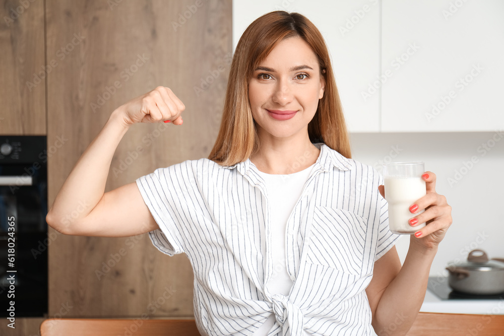 Young woman with glass of milk showing muscles in kitchen