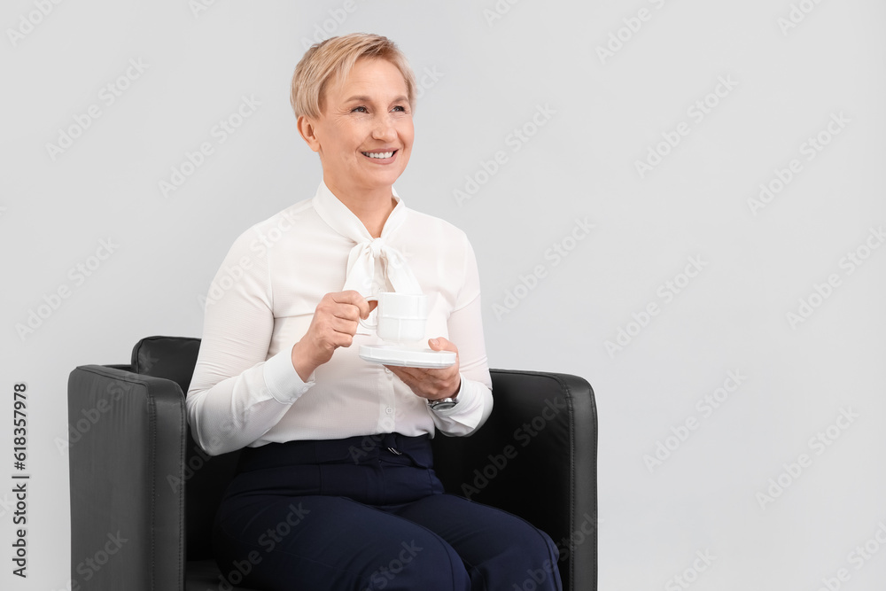 Mature businesswoman drinking coffee while sitting in armchair against grey background