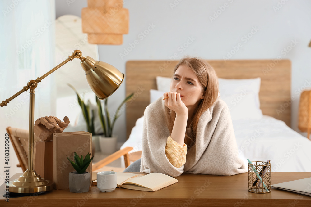 Thoughtful young woman with notebook at table in bedroom