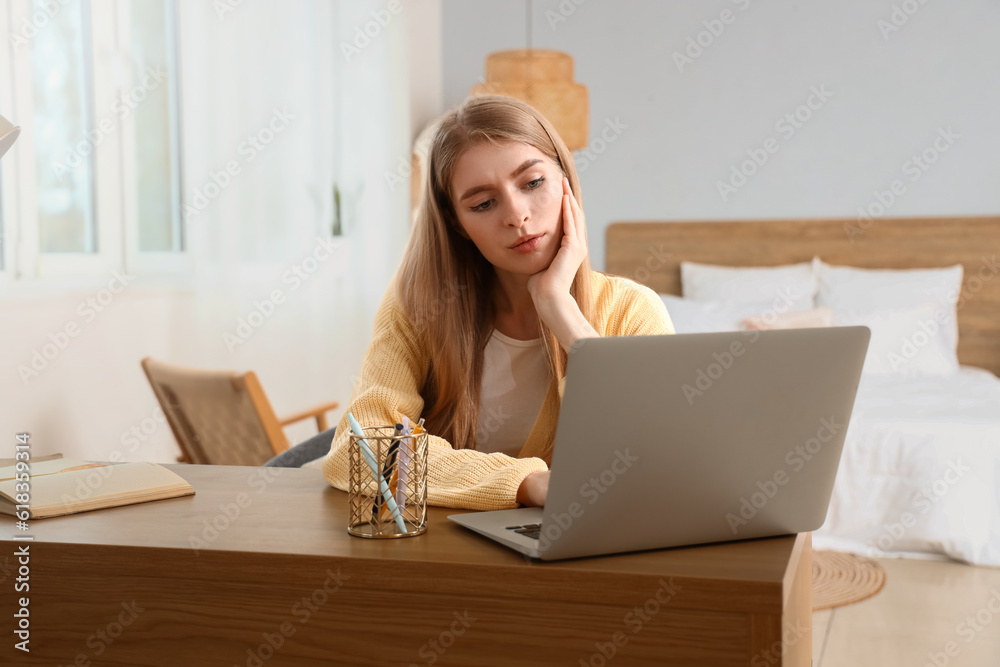 Thoughtful young woman with laptop at table in bedroom