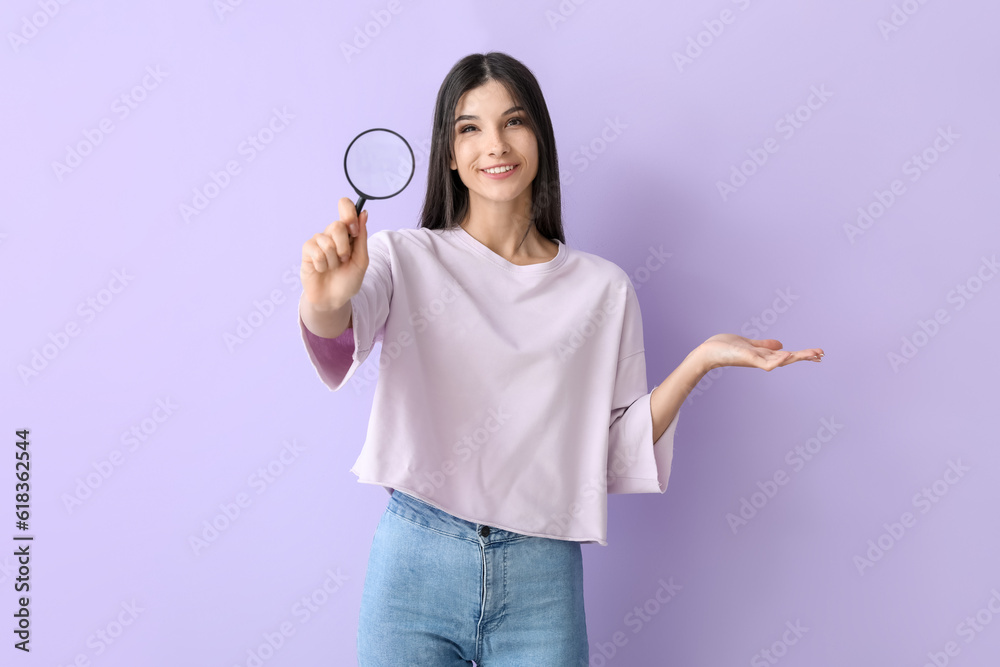 Beautiful happy young woman with magnifier pointing at something on lilac background