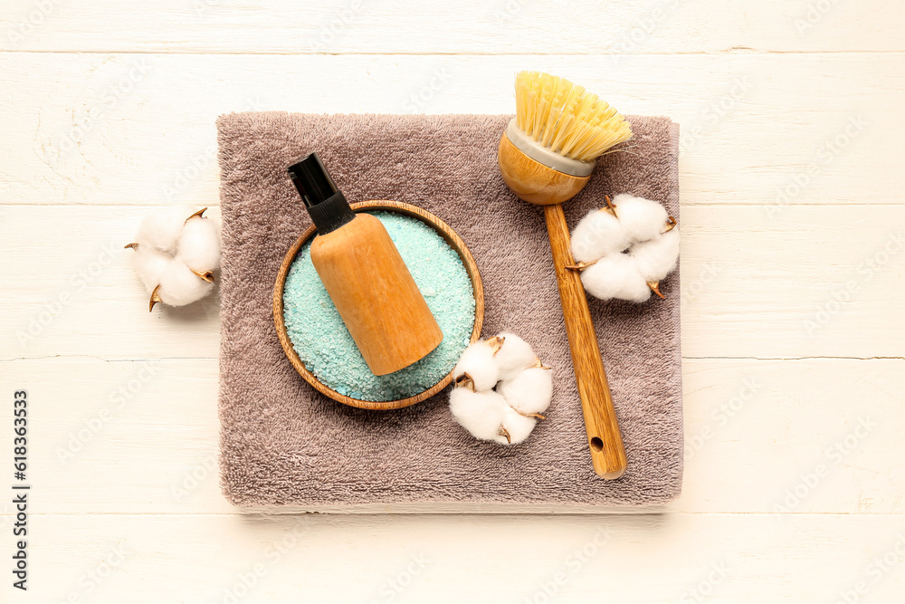 Bottle of cosmetic product, sea salt, massage brush, clean towel and cotton flowers on light wooden 