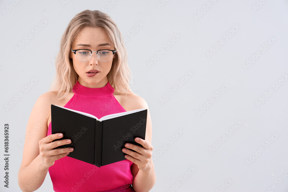 Shocked young woman in stylish eyeglasses reading book on light background