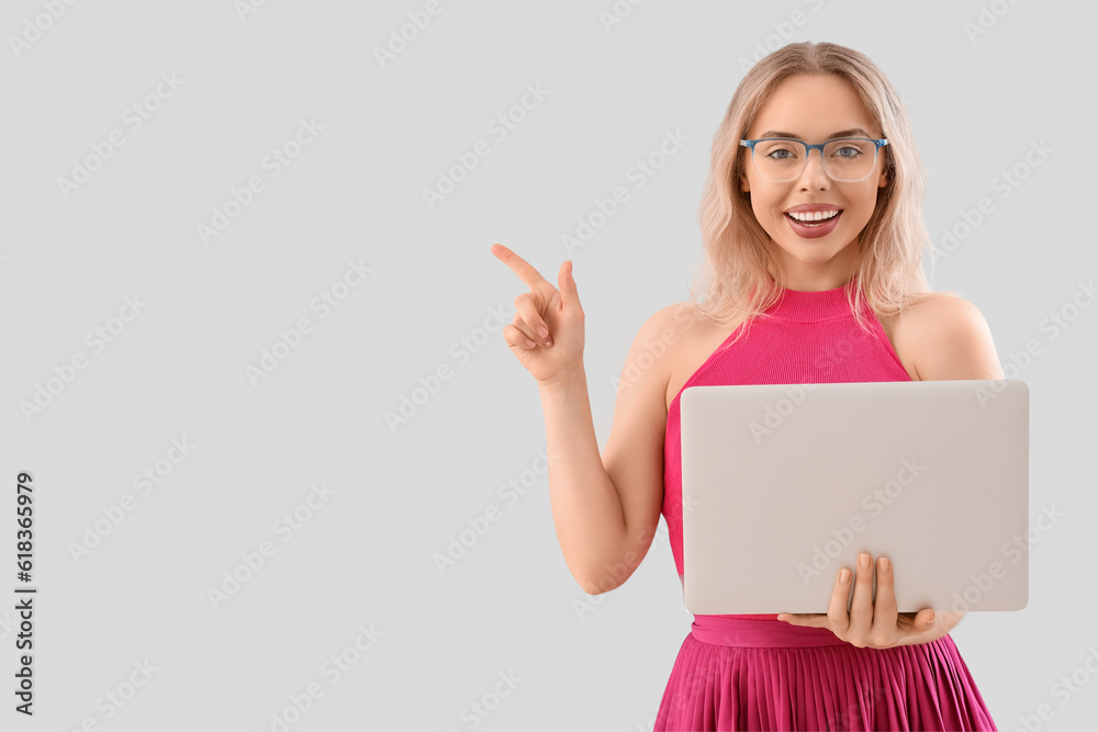 Young woman in stylish eyeglasses with laptop pointing at something on light background