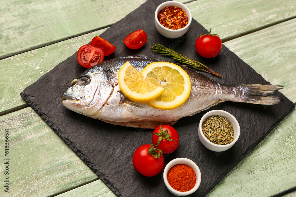 Slate board of raw dorado fish with lemon, tomatoes and spices on green wooden background