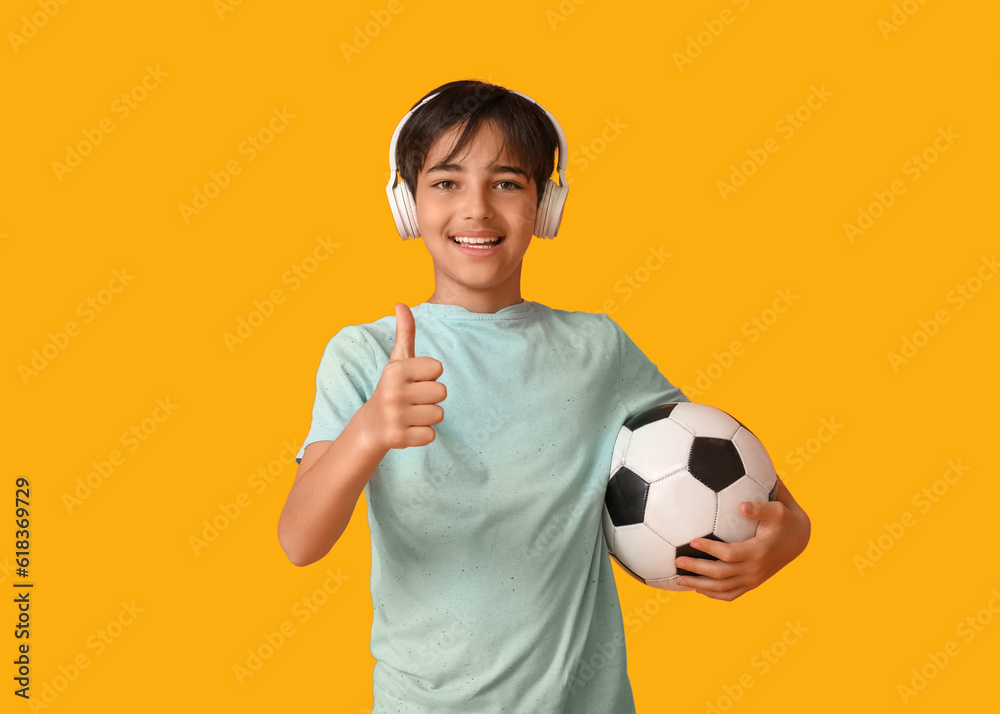 Little boy in headphones with soccer ball showing thumb-up on orange background