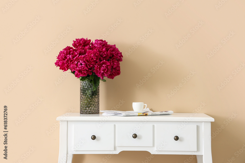 Vase of red peonies with cup of coffee and newspaper on dressing table near beige wall