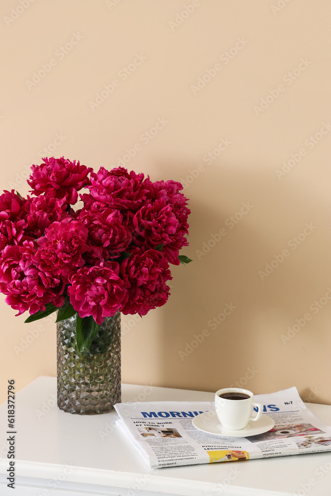 Vase of red peonies with cup of coffee and newspaper on dressing table near beige wall