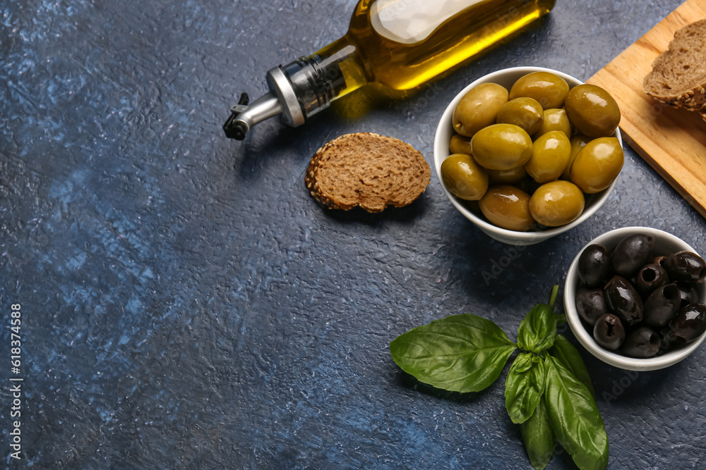 Bowls with ripe olives, bread and bottle of oil on blue background