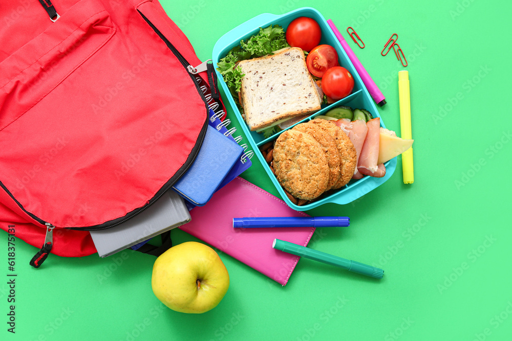 Backpack, stationery and lunch box with tasty food on green background