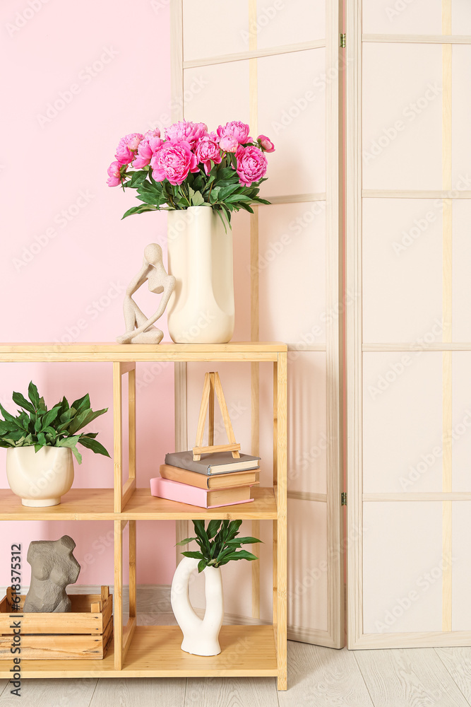 Vase of pink peonies and figurine on shelving unit with dressing screen near color wall