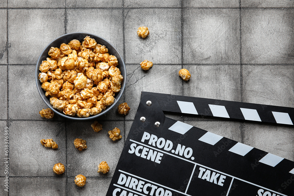 Bowl with tasty popcorn and clapperboard on grey tile background