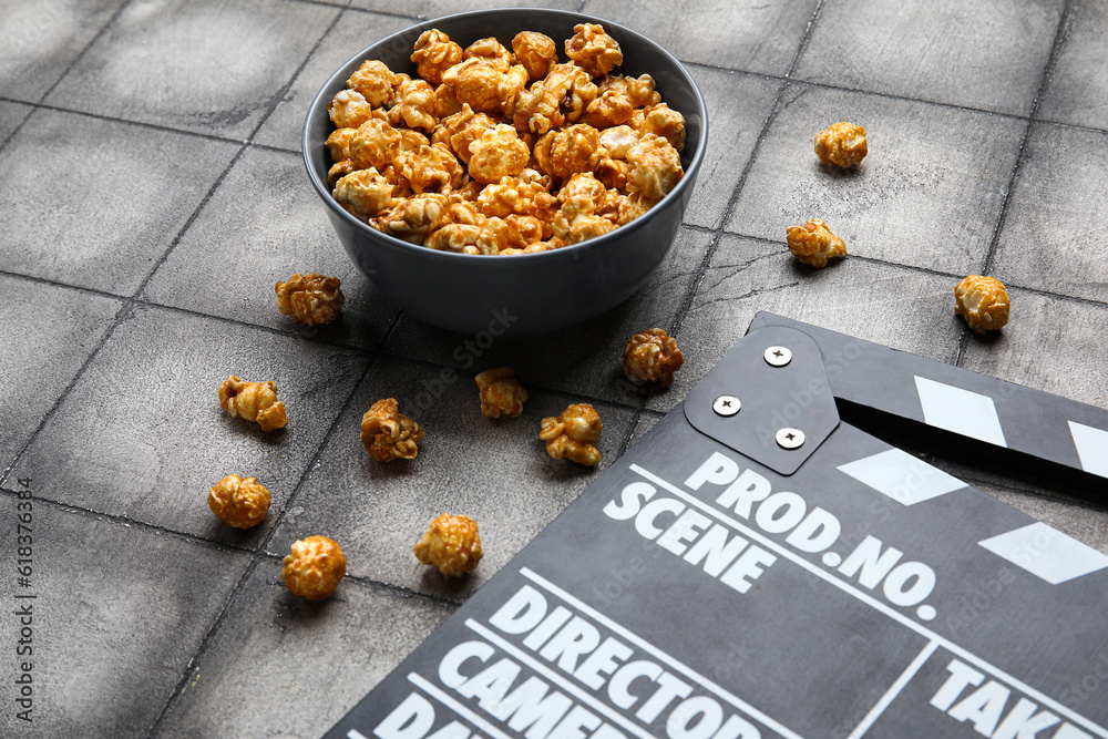 Bowl with tasty popcorn and clapperboard on grey tile background