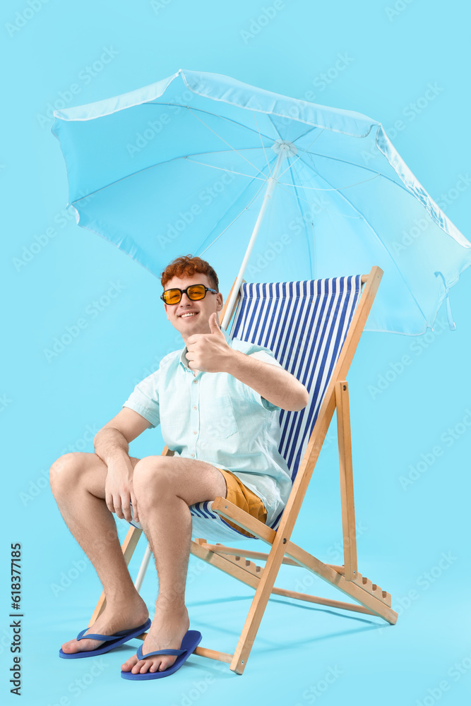 Young man showing thumb-up in deck chair on blue background