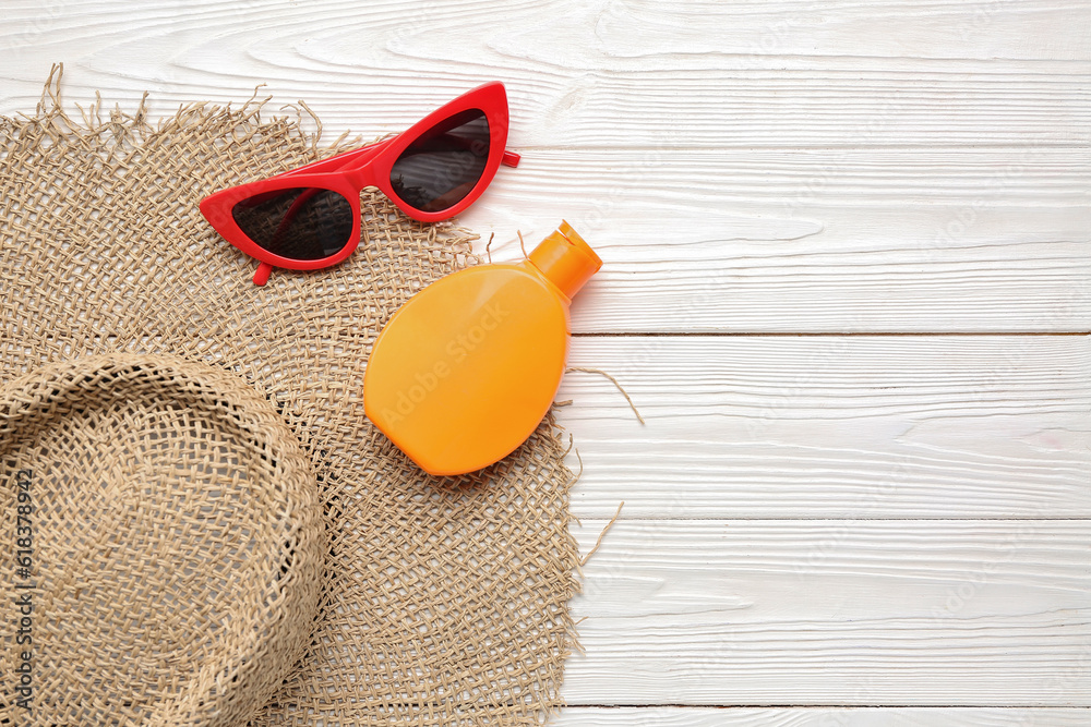 Wicker hat with sunglasses and bottle of sunscreen cream on white wooden background