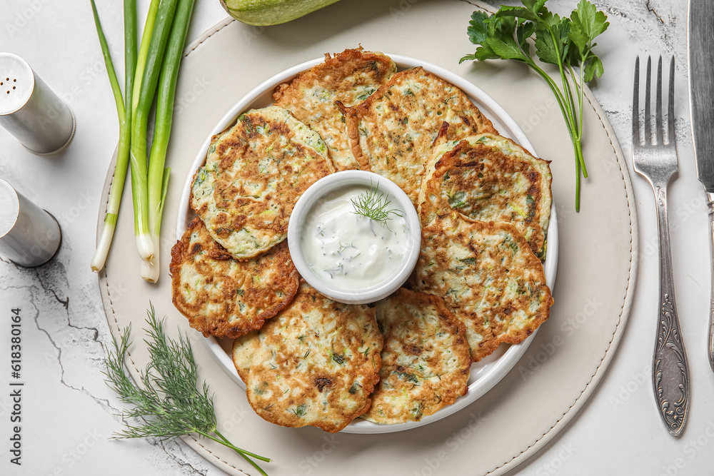 Plate of tasty zucchini fritters with sour cream on white marble background