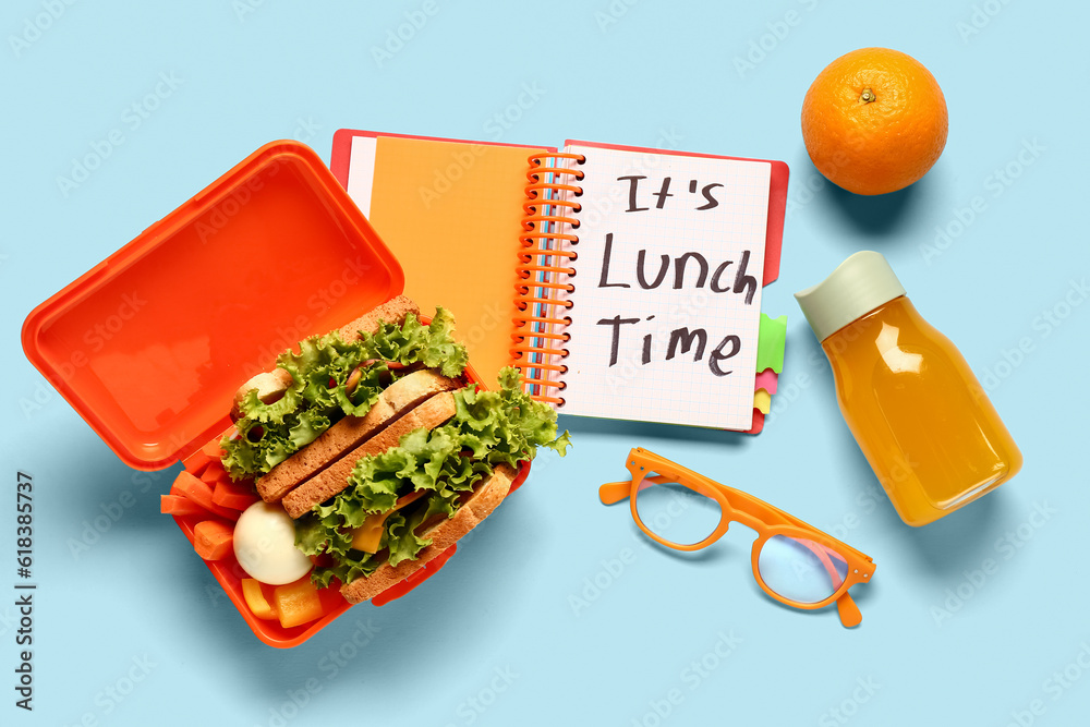 Notebook with text ITS LUNCH TIME, eyeglasses and tasty food in lunchbox on blue background