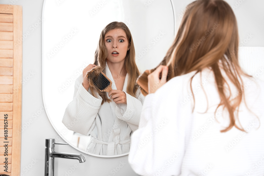 Shocked young woman with hair loss problem brushing near mirror in bathroom