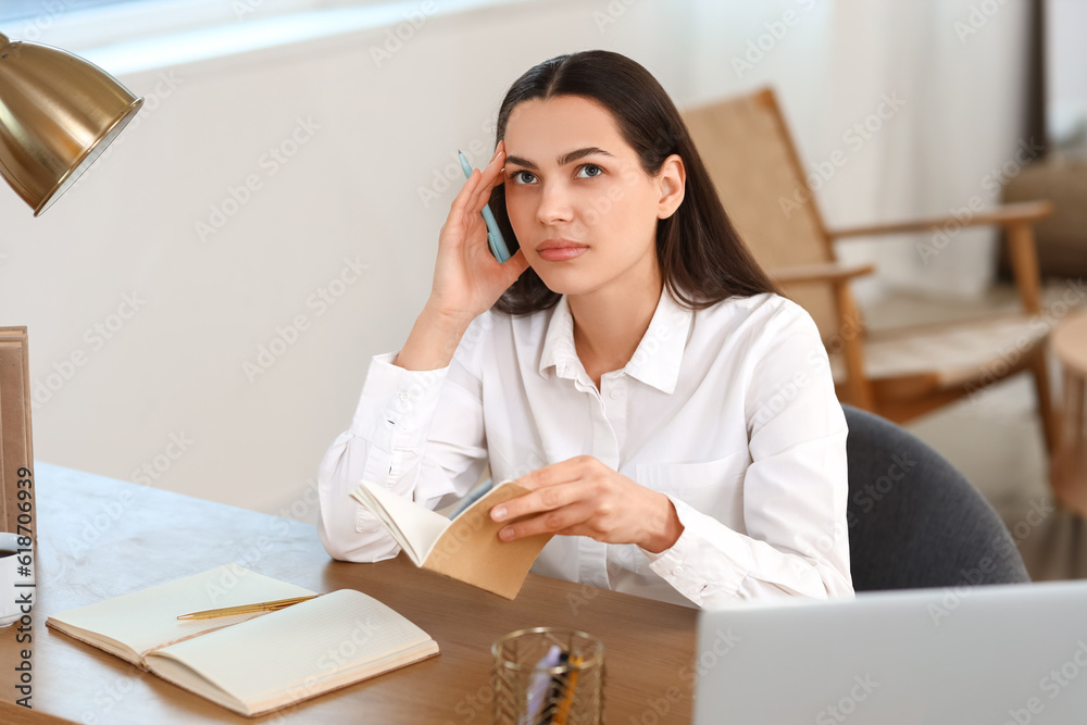 Thoughtful young woman with notebook sitting at table in bedroom