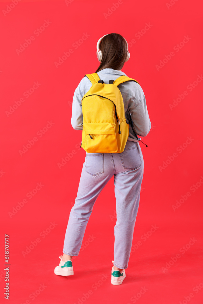 Female student in headphones with backpack on red background, back view