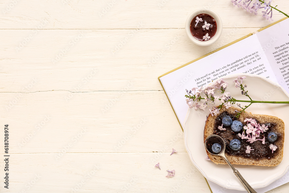 Opened book, plate with sweet jam toast and flowers on white wooden table